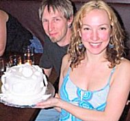 Jenna is smiling a huge bright grin in the forefront. Next to her is a white anniversary cake with icing seashells. Behind her is her husband Chris, a cute guy with an impish grin and a soulpatch.below his lower lip to his chin,.