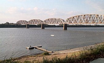 This image shows an long old, rusty train bridge spanning a wide river with a green bank on the other side and a small dock below.